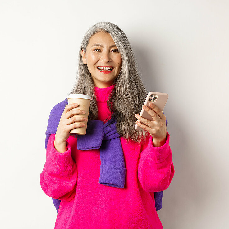 Sujet_Frau_Smartphone_Technik_Handy E-commerce concept. Beautiful asian senior woman drinking coffee from takeaway cup and using mobile phone, smiling at camera, white background